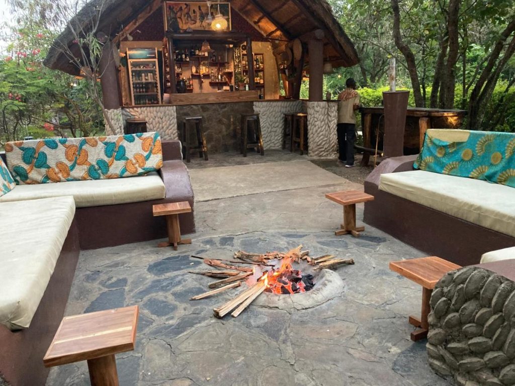 A cozy outdoor lounge at Camp Bastian Mikumi featuring a central stone fire pit, built-in sofas with colorful African-print cushions, and a thatched-roof bar in the background.
