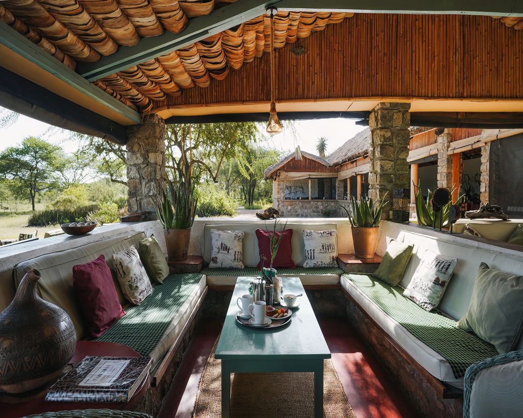 : Ndutu cottages' central seating area with low-slung, cushioned stone benches, colorful throw pillows, and a wooden coffee table set for tea under a thatched and bamboo-lined roof.