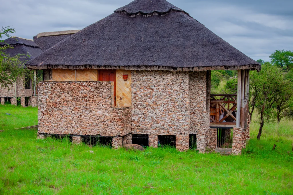 Exterior of a traditional stone and thatched-roof luxury villa at Serengeti Safari Lodge in Tanzania, featuring a private wooden balcony and elevated foundation on a green savannah landscape.