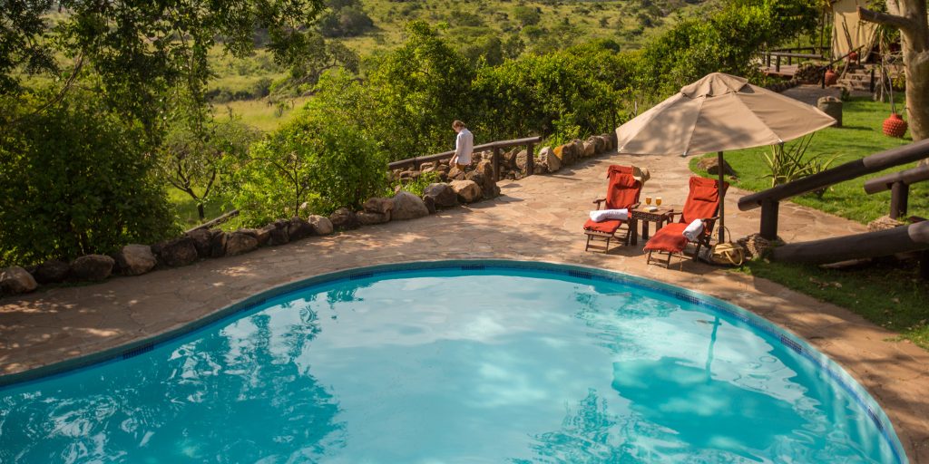 A round turquoise swimming pool on a stone terrace at Safari Haven Migration Camp, featuring red lounge chairs under a tan umbrella with a panoramic view of the lush Serengeti valley.