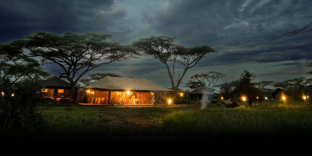 large, illuminated canvas safari tent under a dark, cloudy twilight sky in the Serengeti, surrounded by acacia trees and glowing lanterns.