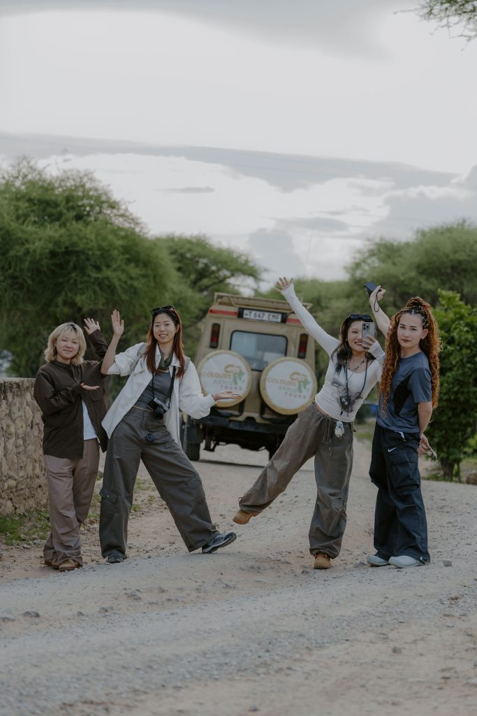 Four women posing playfully on a dirt road in front of a beige Tanzania Safari Makers vehicle during a guided tour.