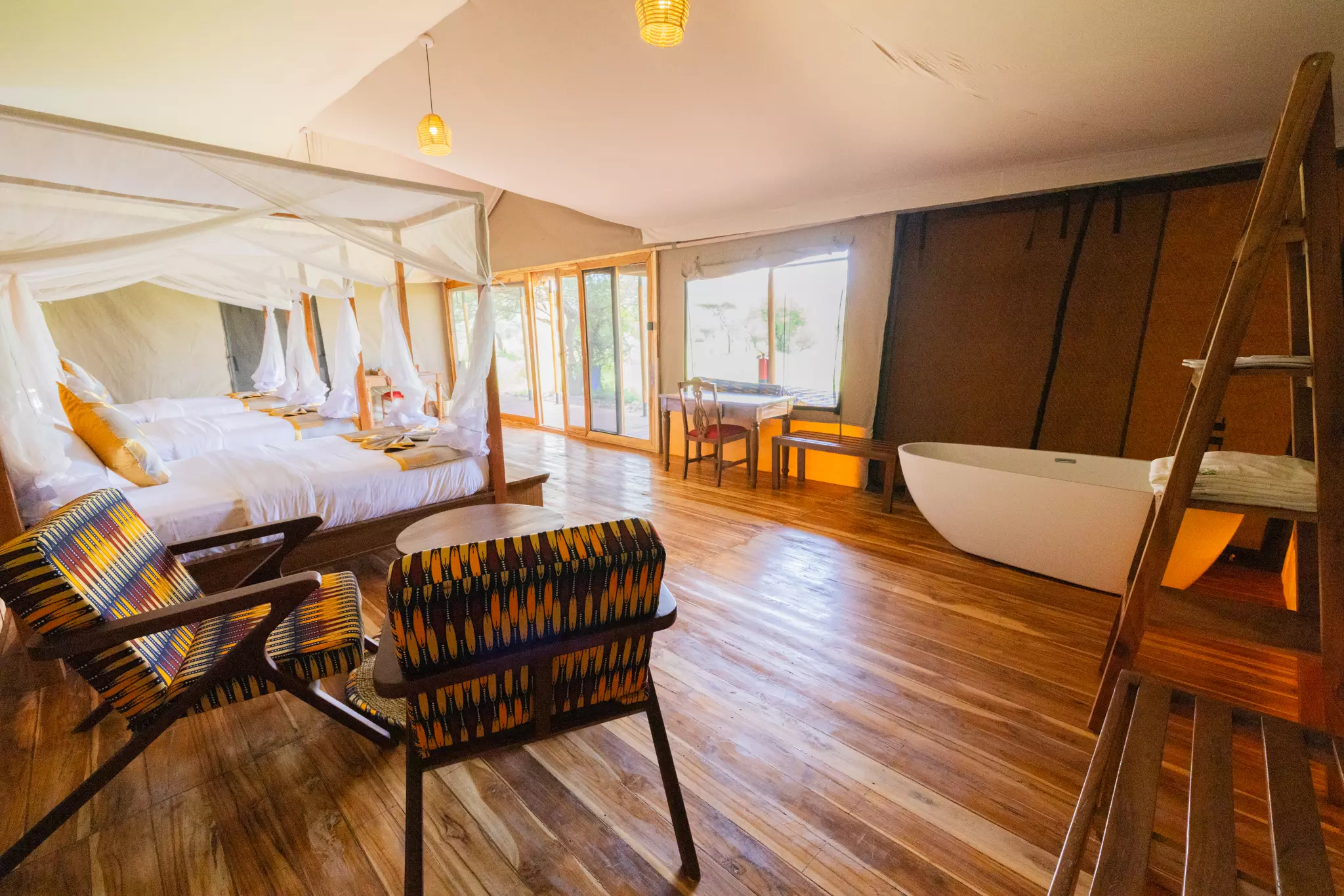 Spacious interior of a luxury safari tent featuring multiple beds with mosquito nets, a freestanding white bathtub, wooden floors, and African-patterned armchairs overlooking the Serengeti.