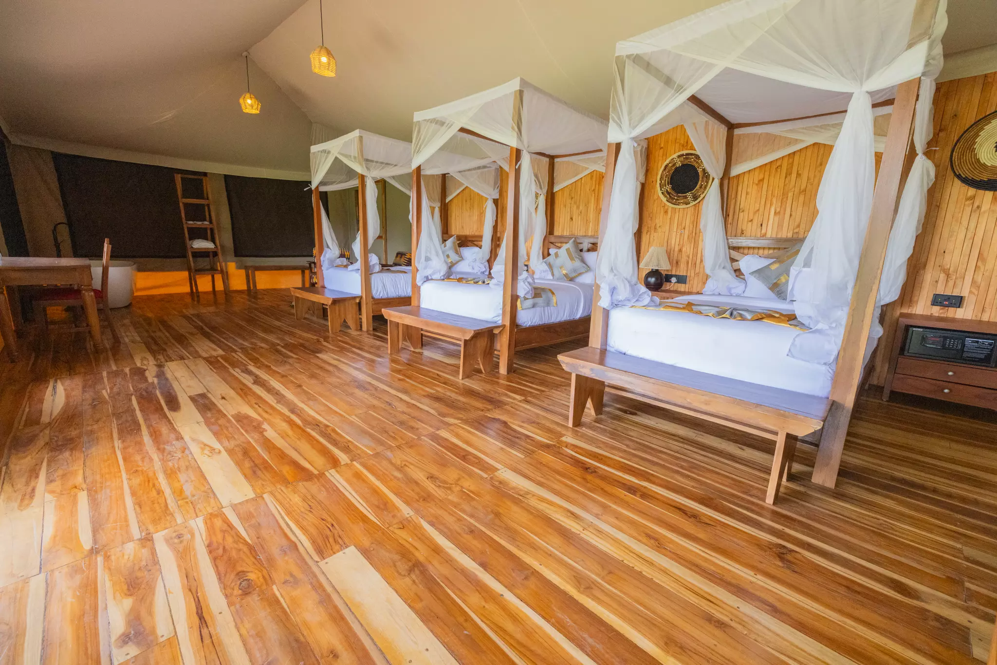 A wide-angle view of a large, luxury safari tent interior with polished light wood flooring, featuring three four-poster wooden beds with white mosquito nets and linens, a small desk, and warm pendant lighting.